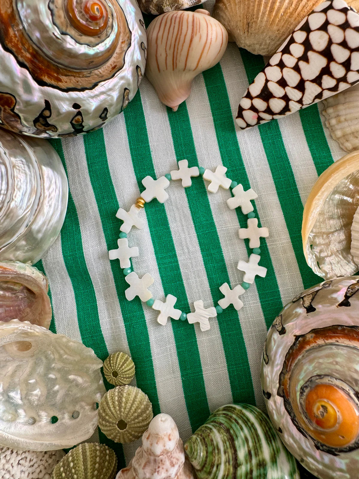 Handcrafted St Jean Bracelet with mother of pearl cross beads, surrounded by seashells on a striped green fabric.