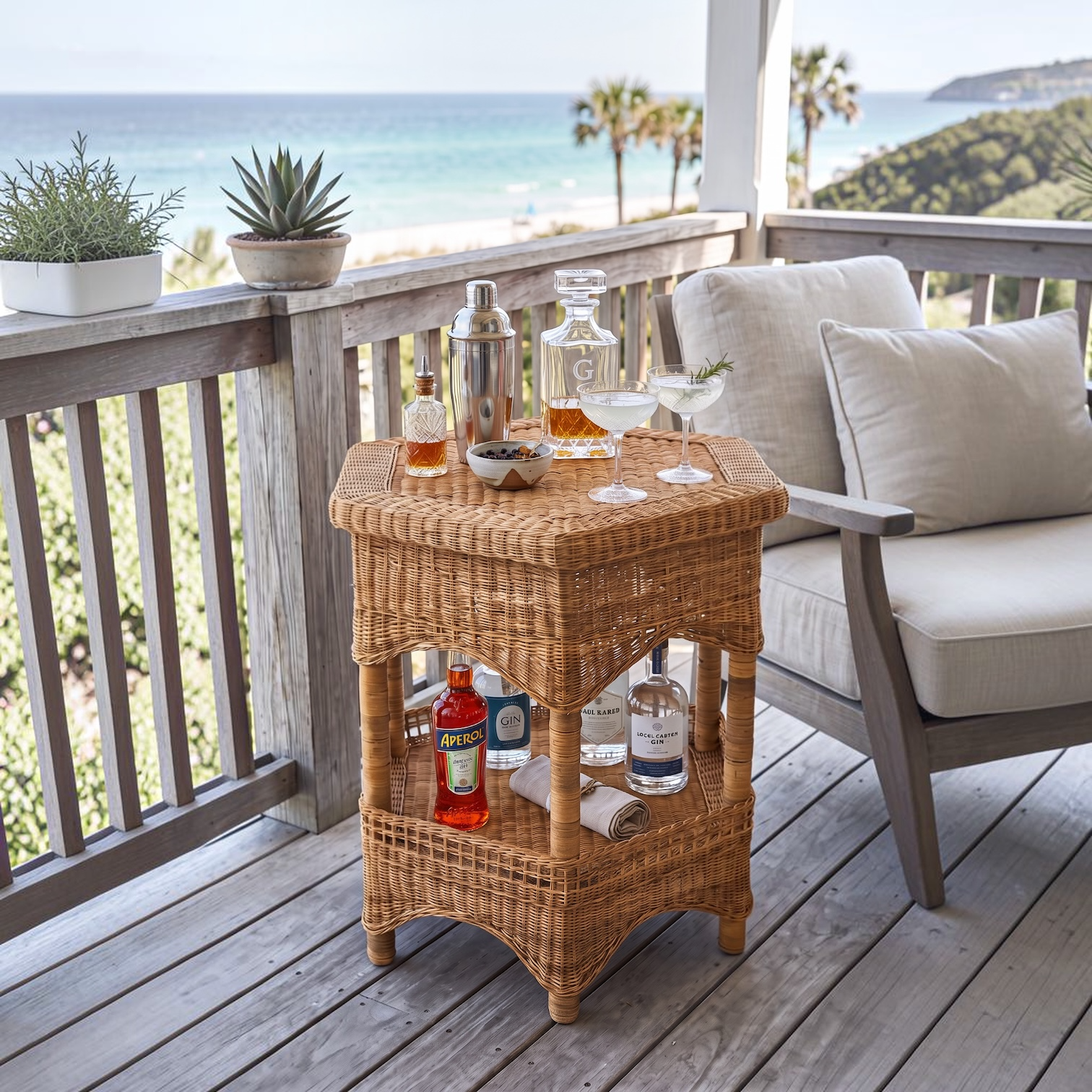 A woven rattan outdoor bar cart on a wooden deck beside a beach chair, styled with bottles, glasses, and a cocktail decanter, with an ocean and palm tree view in the background.
