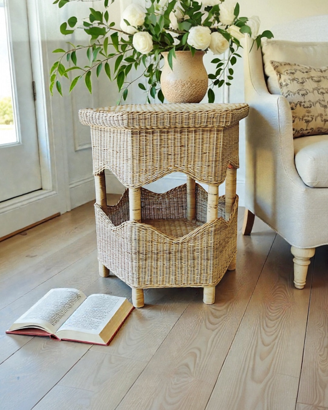 Wicker side table with a vase of flowers and an open book on a wooden floor.