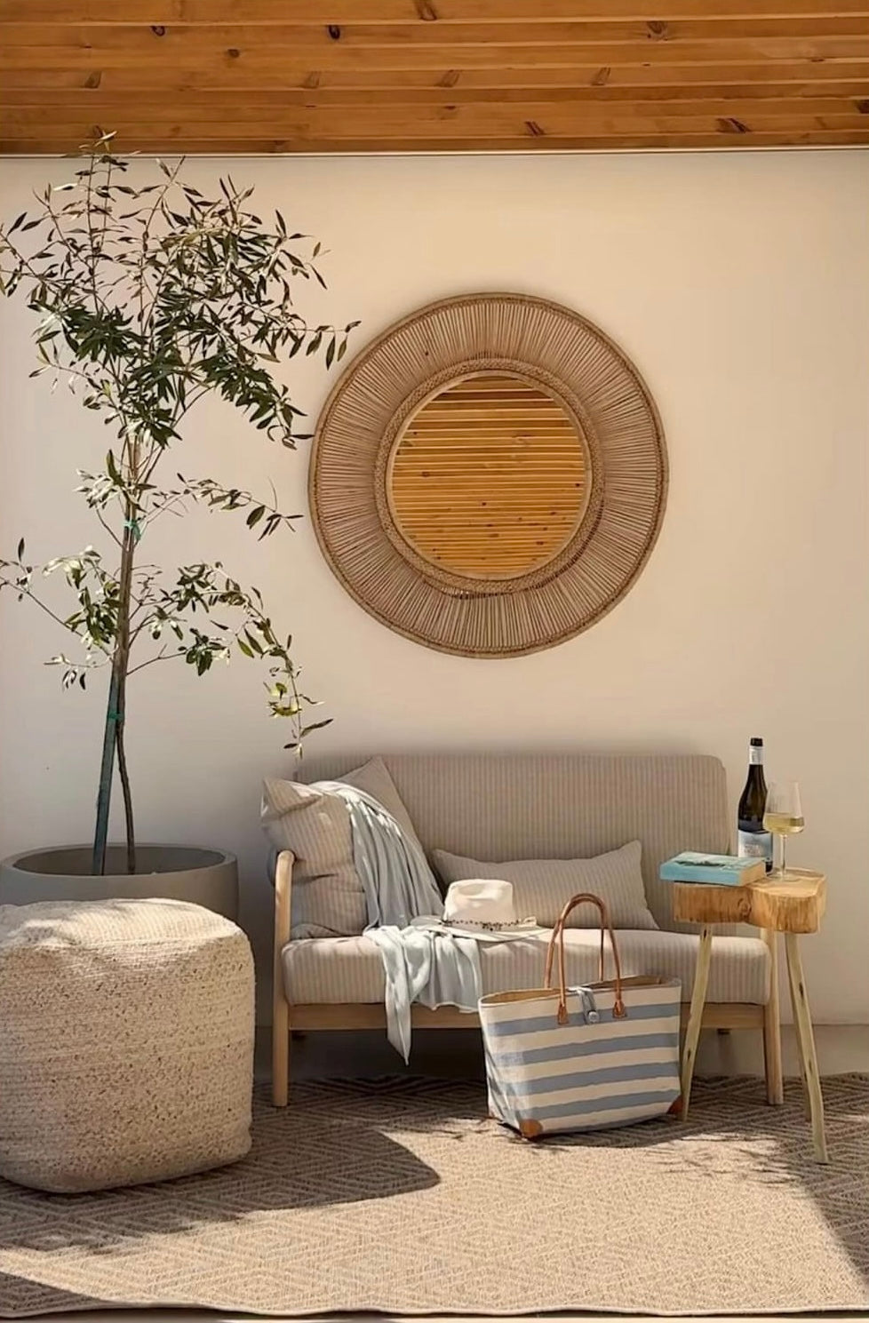 Handwoven cane mirror above a cozy sofa with a striped beach shopper bag, plant, and a wooden table in a bright living space.