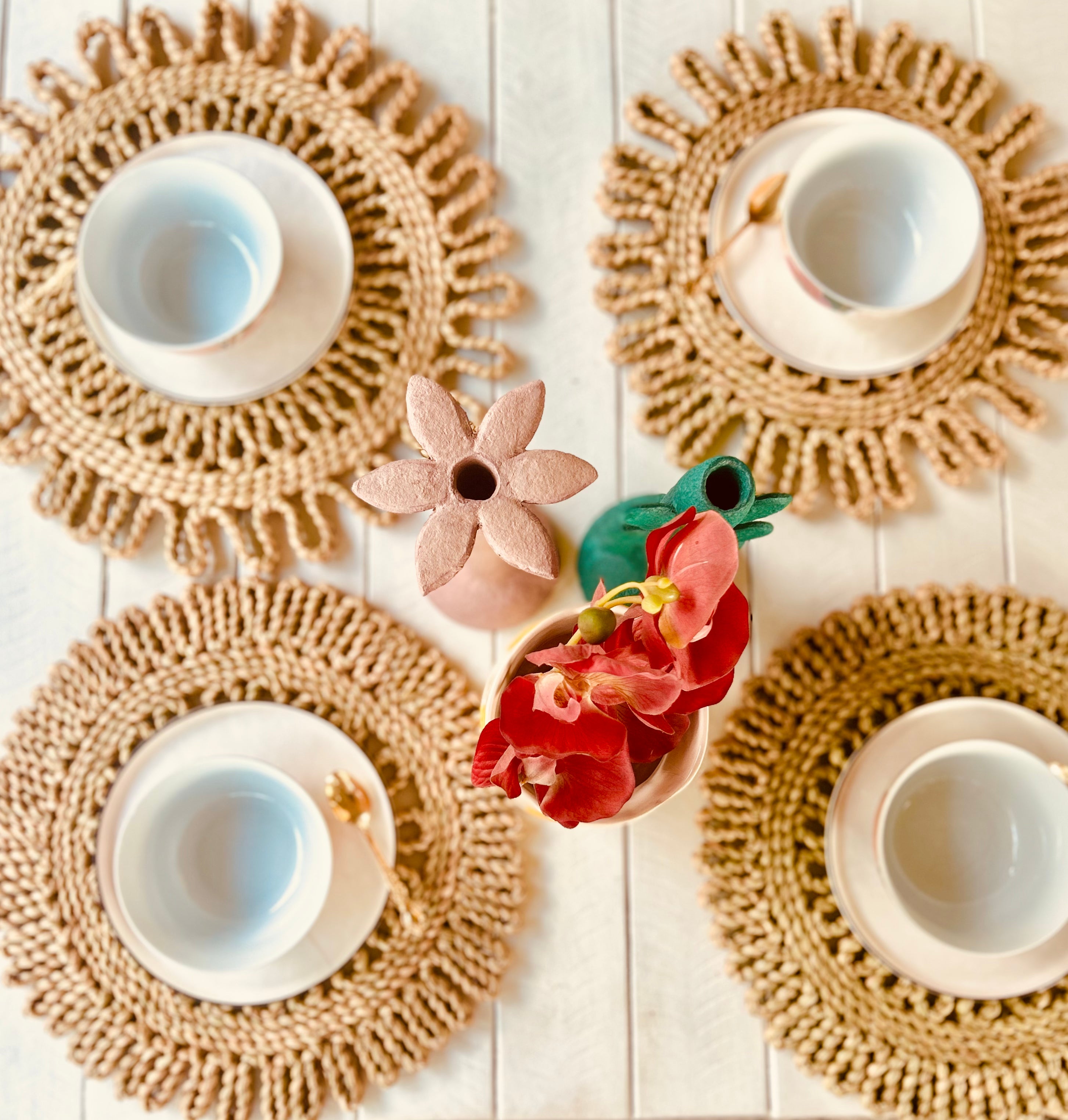 Handwoven scalloped placemats with white cups and vibrant flowers on a wooden table setting. Perfect for any dining occasion.