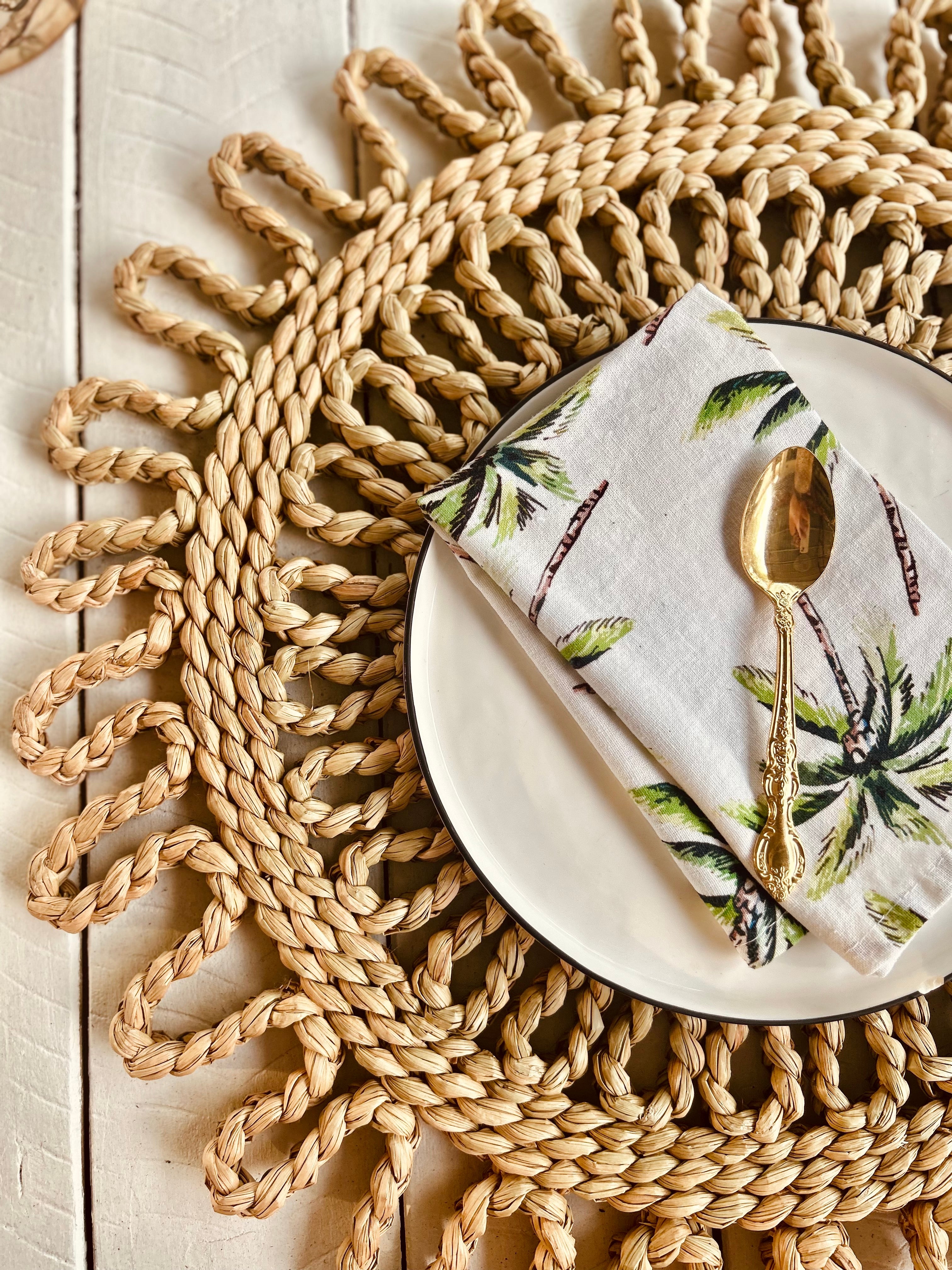 Handwoven scalloped placemat with a white plate, tropical napkin, and gold spoon, perfect for dining table settings.