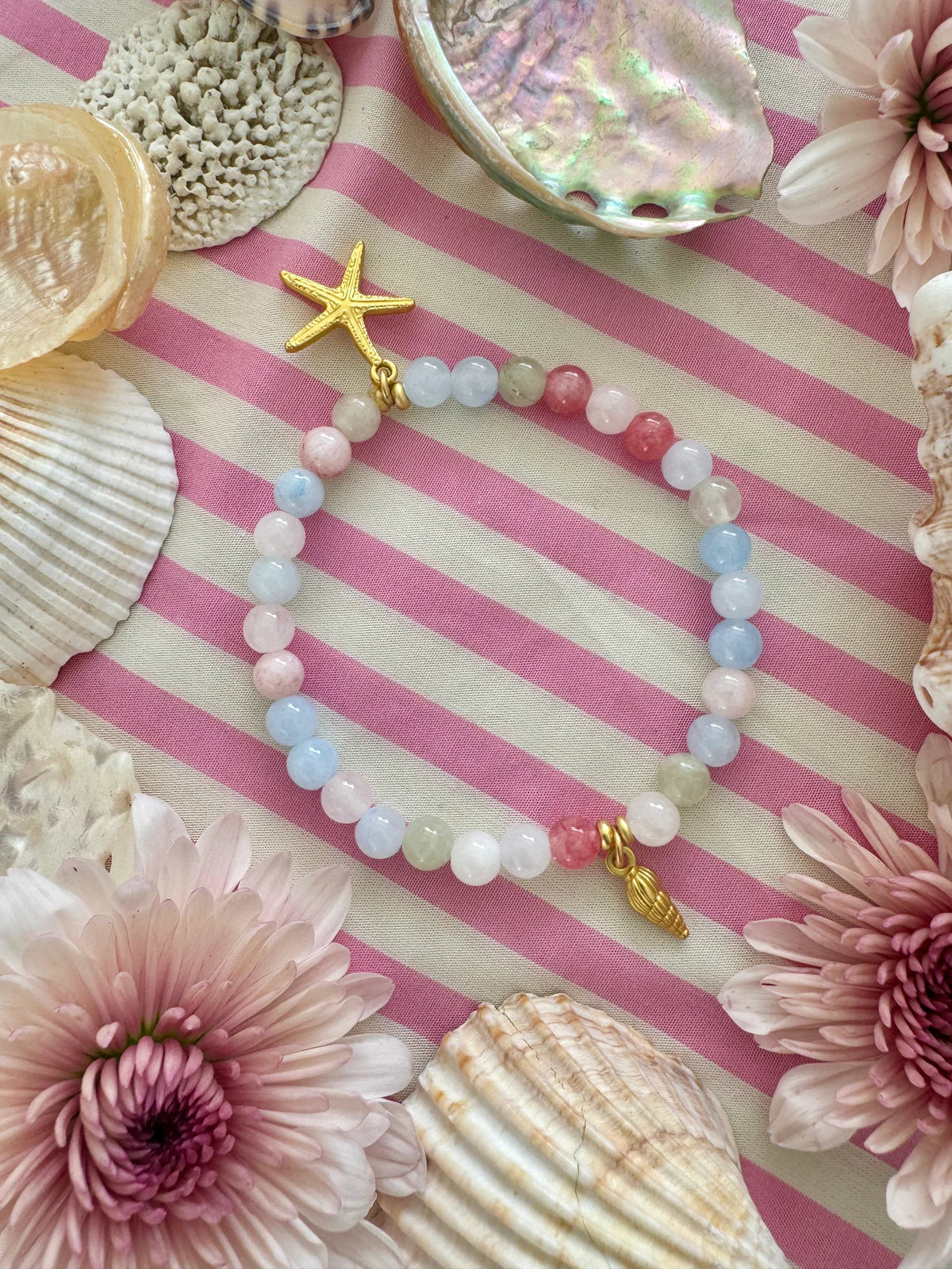 Handcrafted ocean inspired Kiki Bracelet with pastel beads and gold charms on a pink striped background.