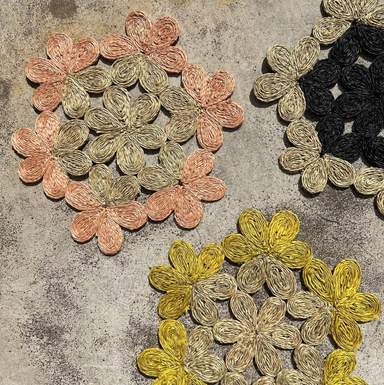 Handwoven floral placemats in pink, yellow, and black on a textured background, perfect for dining or picnics.
