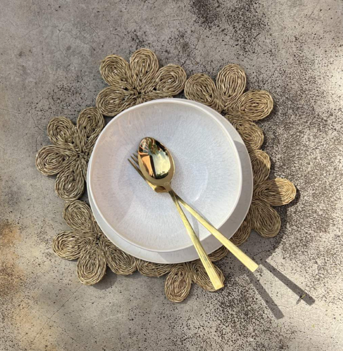 Handwoven floral-inspired placemat with a white plate and gold cutlery on a textured surface.