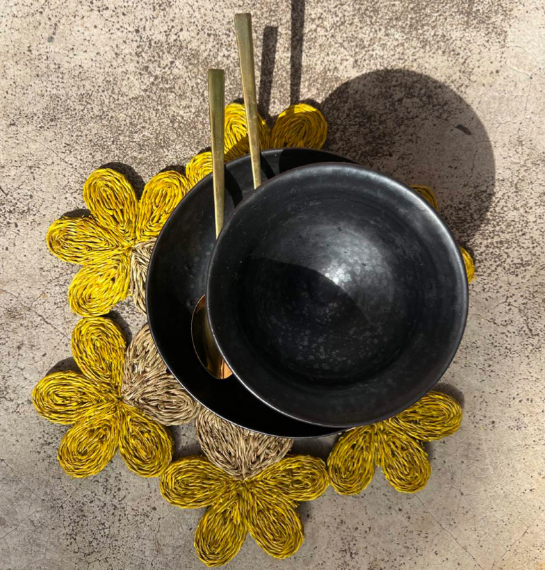 Handwoven floral placemat in yellow with black bowls and gold utensils, perfect for dining or beach picnics.