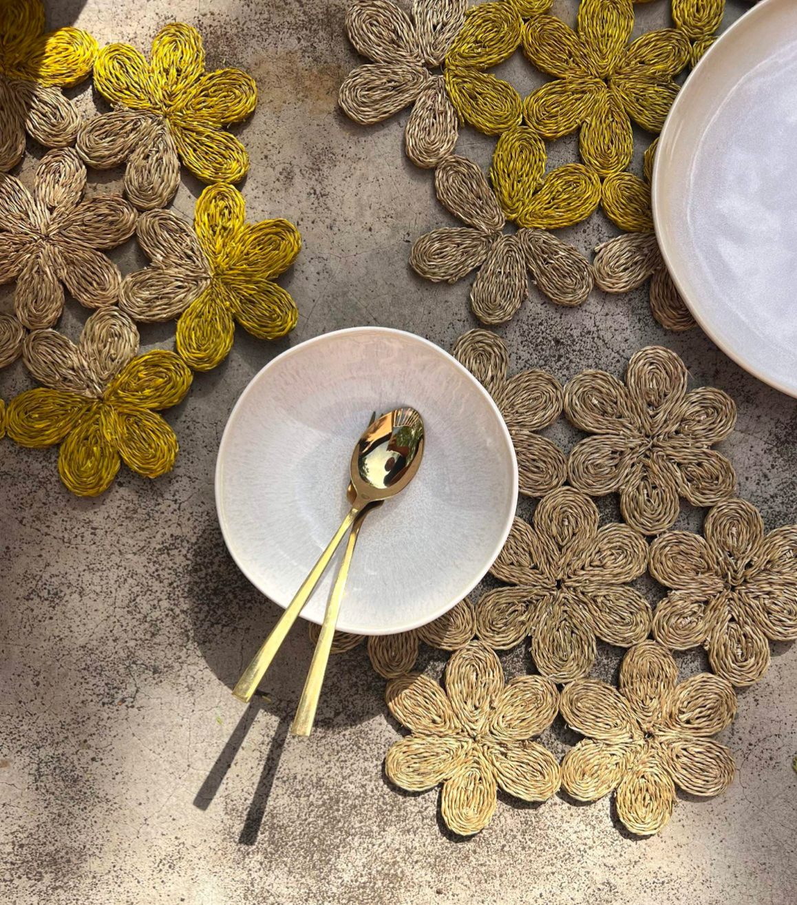 Handwoven floral inspired placemats in yellow and natural tones with a white bowl and golden utensils on a textured surface.