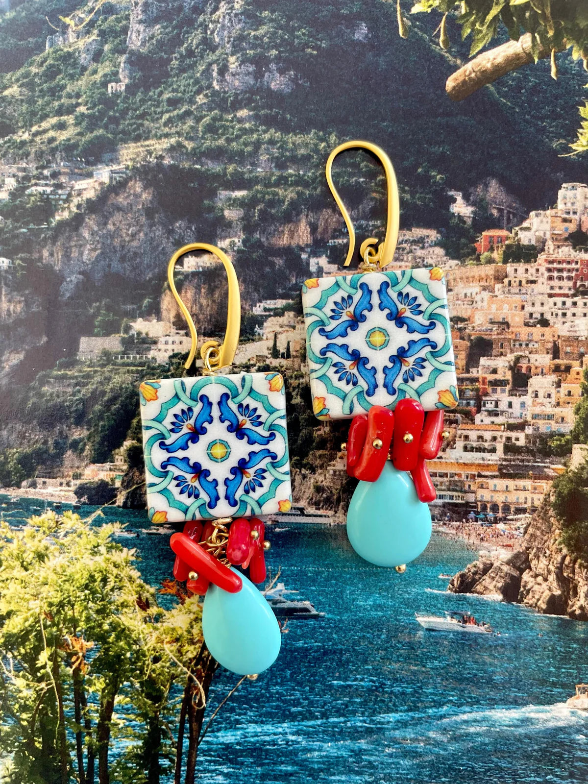 Handcrafted ocean inspired Del Vento earrings with ceramic tile, turquoise drop, and coral charms against a Mediterranean background.