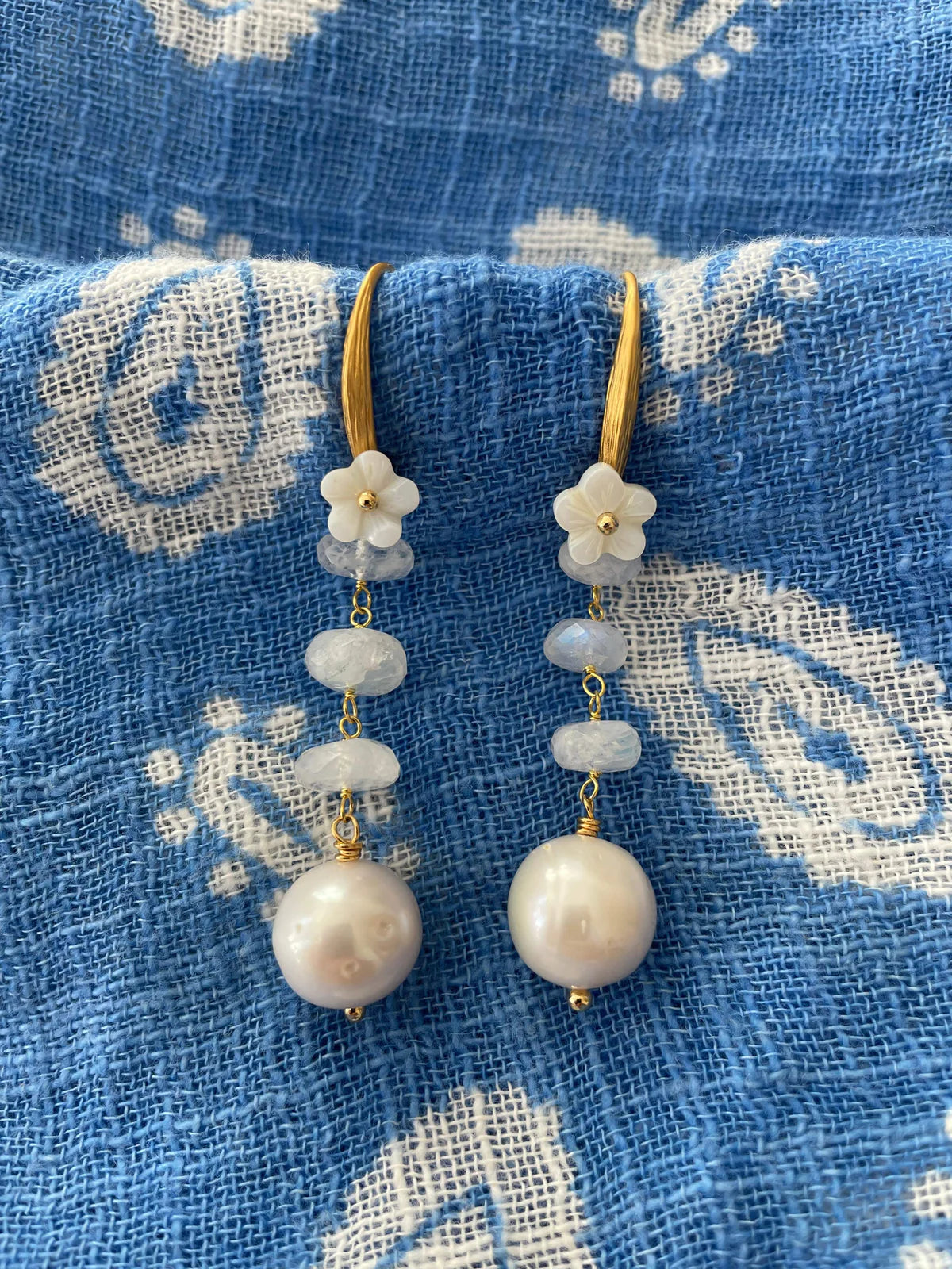 Handcrafted Gabriella Earrings featuring unique baroque pearls, mother of pearl flowers, and moonstone beads on a blue textile background.