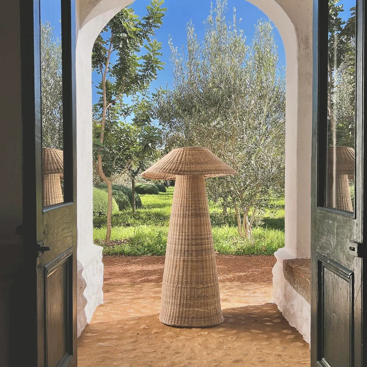 Intaba Cane Woven Floor Lamp standing in an archway overlooking a lush garden, showcasing its elegant design and natural materials.
