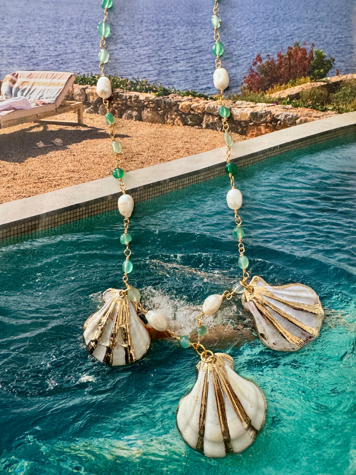 Handcrafted ocean-inspired Sapore di Mare Necklace with gold scallop shells, jade, and pearl, set against a serene pool backdrop.
