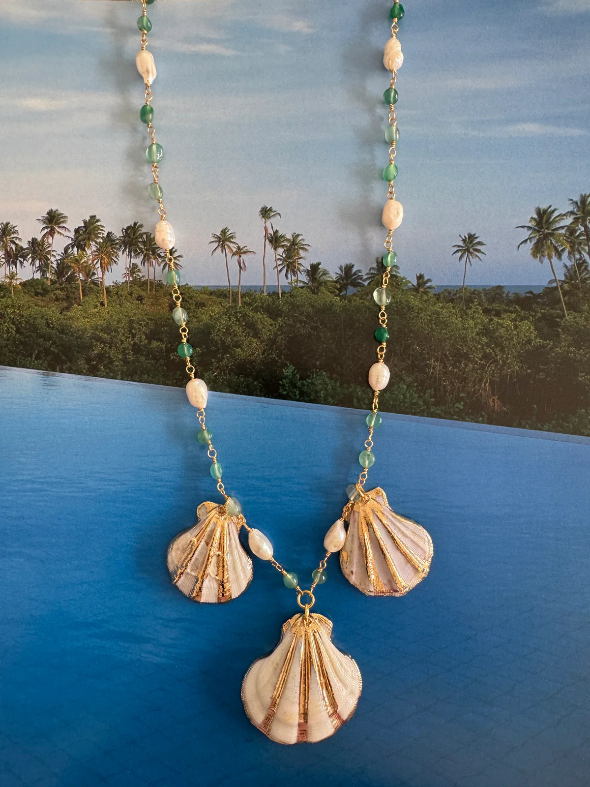 Handcrafted ocean-inspired Sapore di Mare Necklace with gold scallop shells and jade pearl rosary chain against a tropical backdrop.