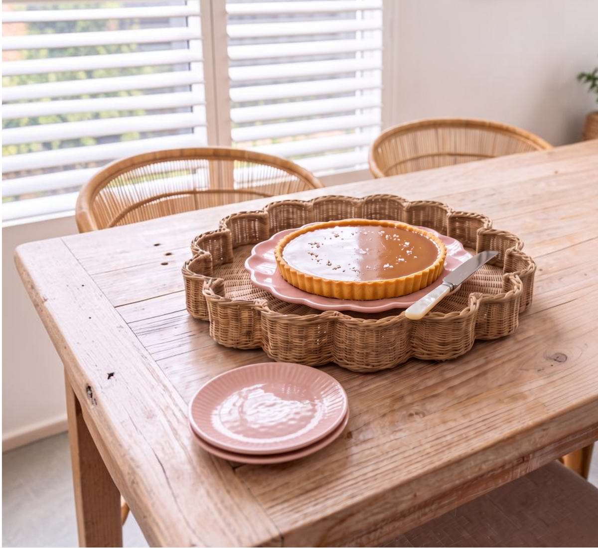A scalloped wicker tray holding a tart and a knife, styled on a wooden dining table with pink plates and wicker chairs in a bright room.