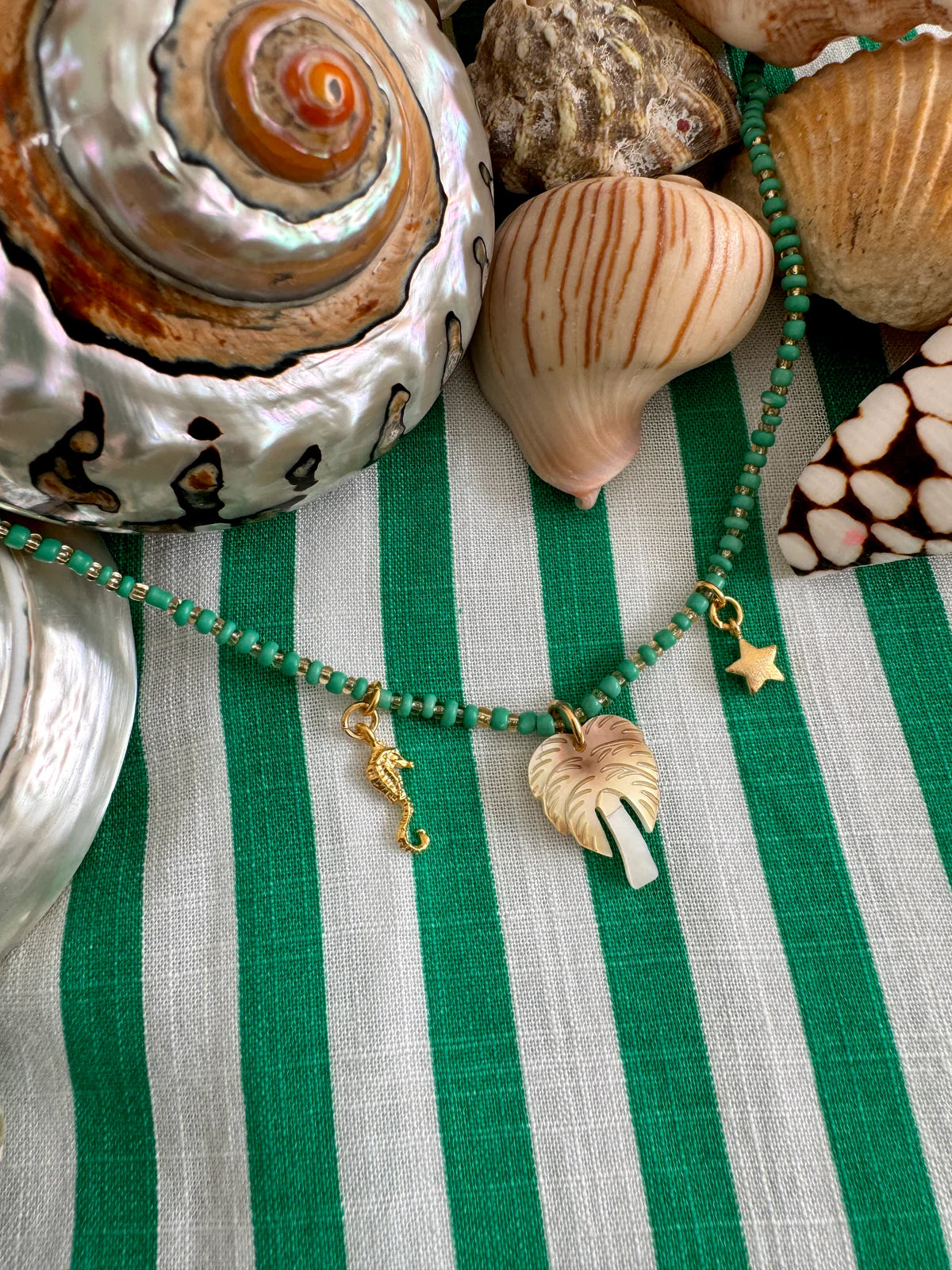 Handcrafted ocean inspired teal necklace with sea horse, palm tree, and star charms on a striped background with seashells.