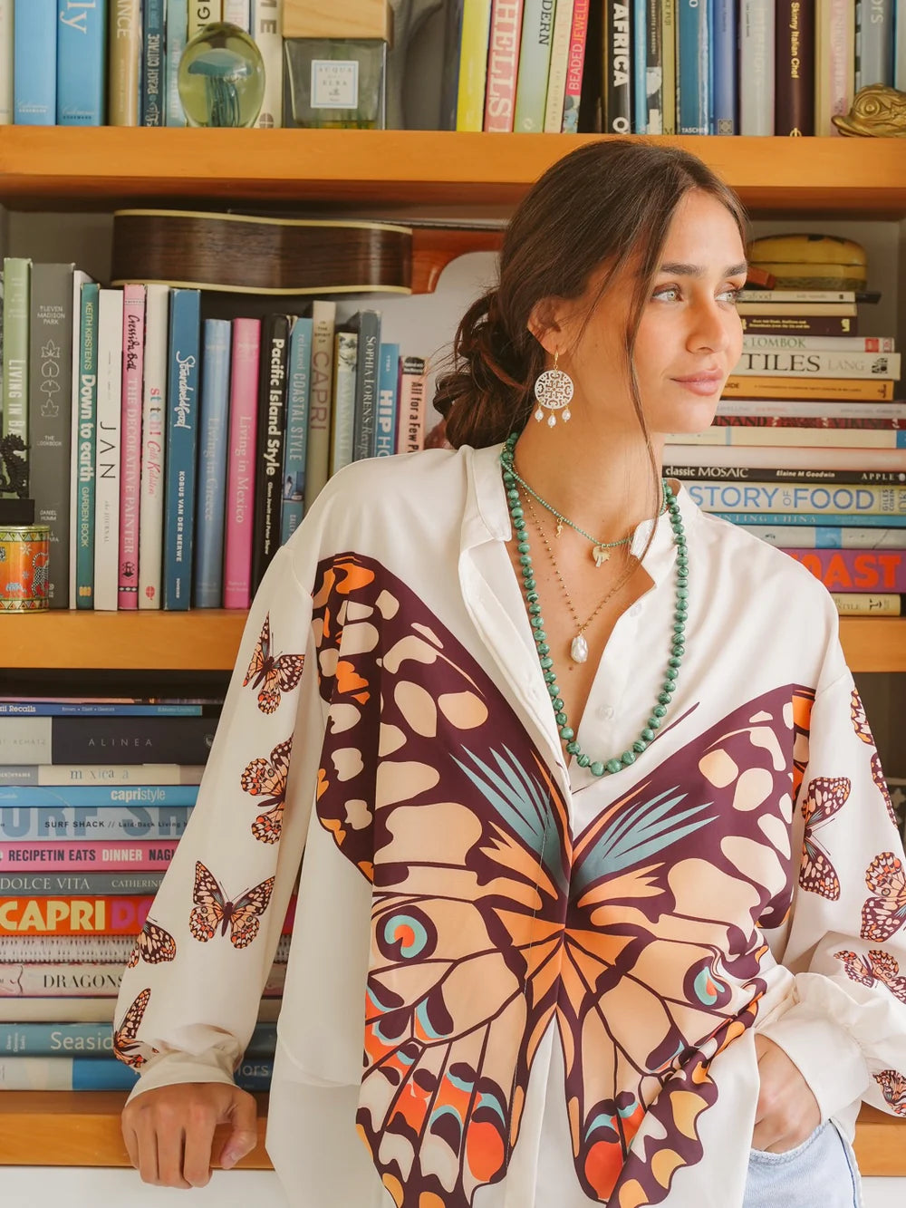 Woman in butterfly print blouse with jewelry, standing near a bookshelf filled with colorful books.