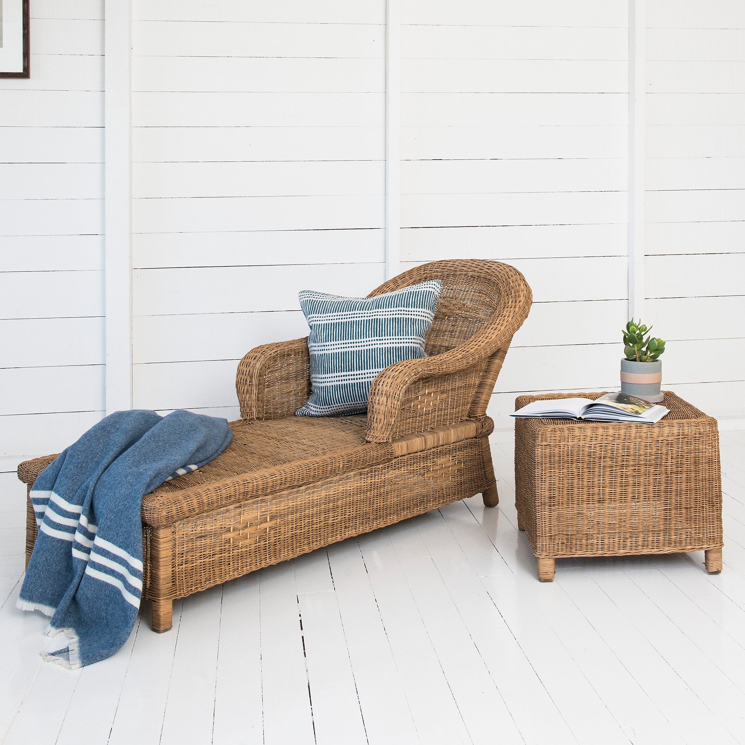 Wicker lounge chair with striped pillow and blue towel against a white wall.