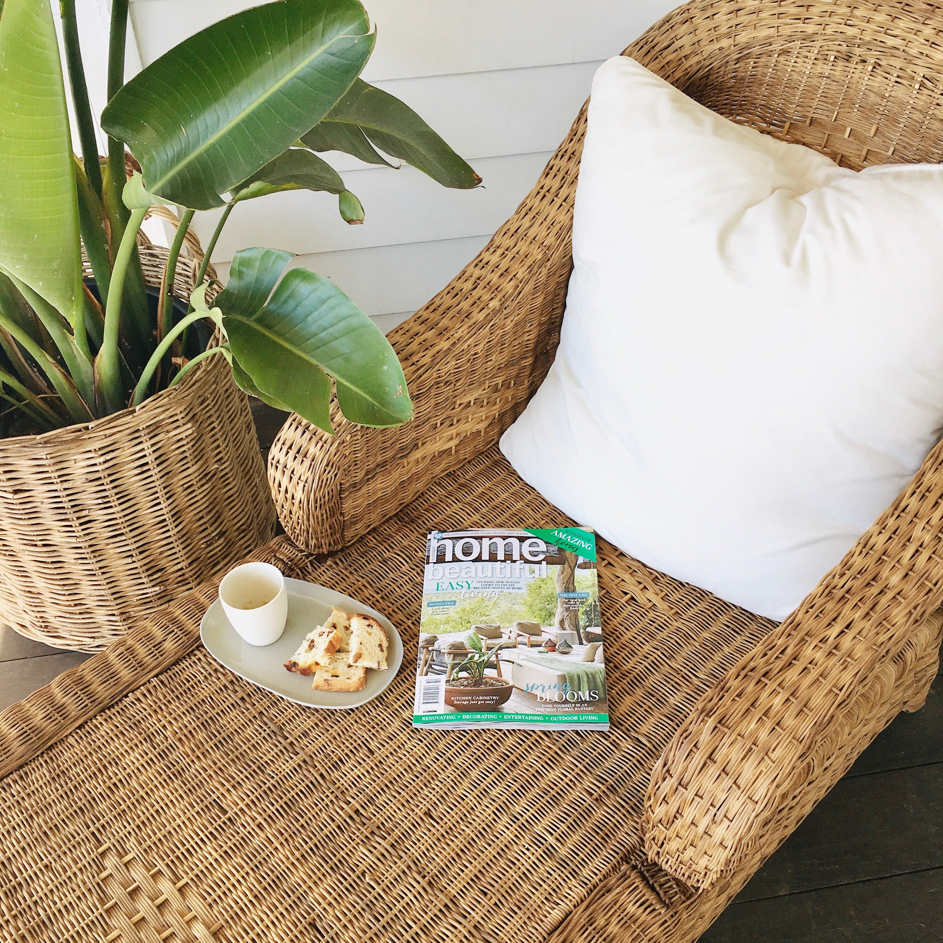 Coastal drift cane lounger with a magazine, cup, and snacks on a wooden floor.