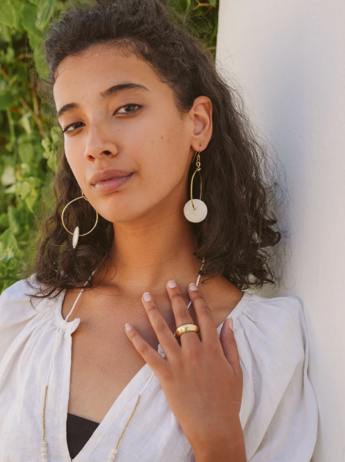 Woman wearing hoop earrings and a ring, standing against a white column with greenery in the background