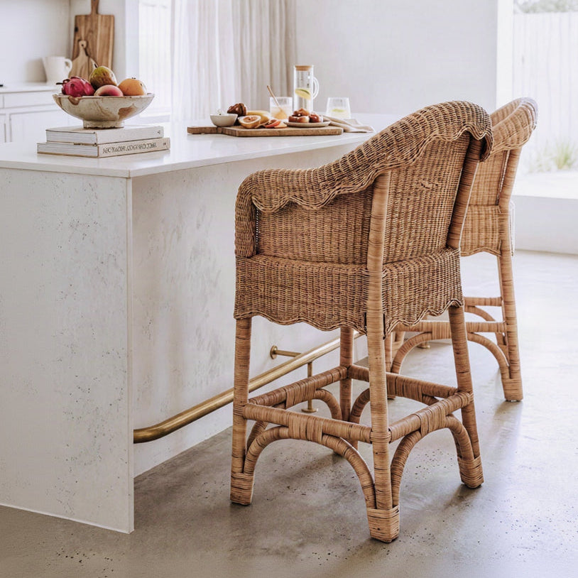 Two natural woven rattan bar stools with scalloped backs, positioned at a white stone kitchen island in a bright, modern room.
