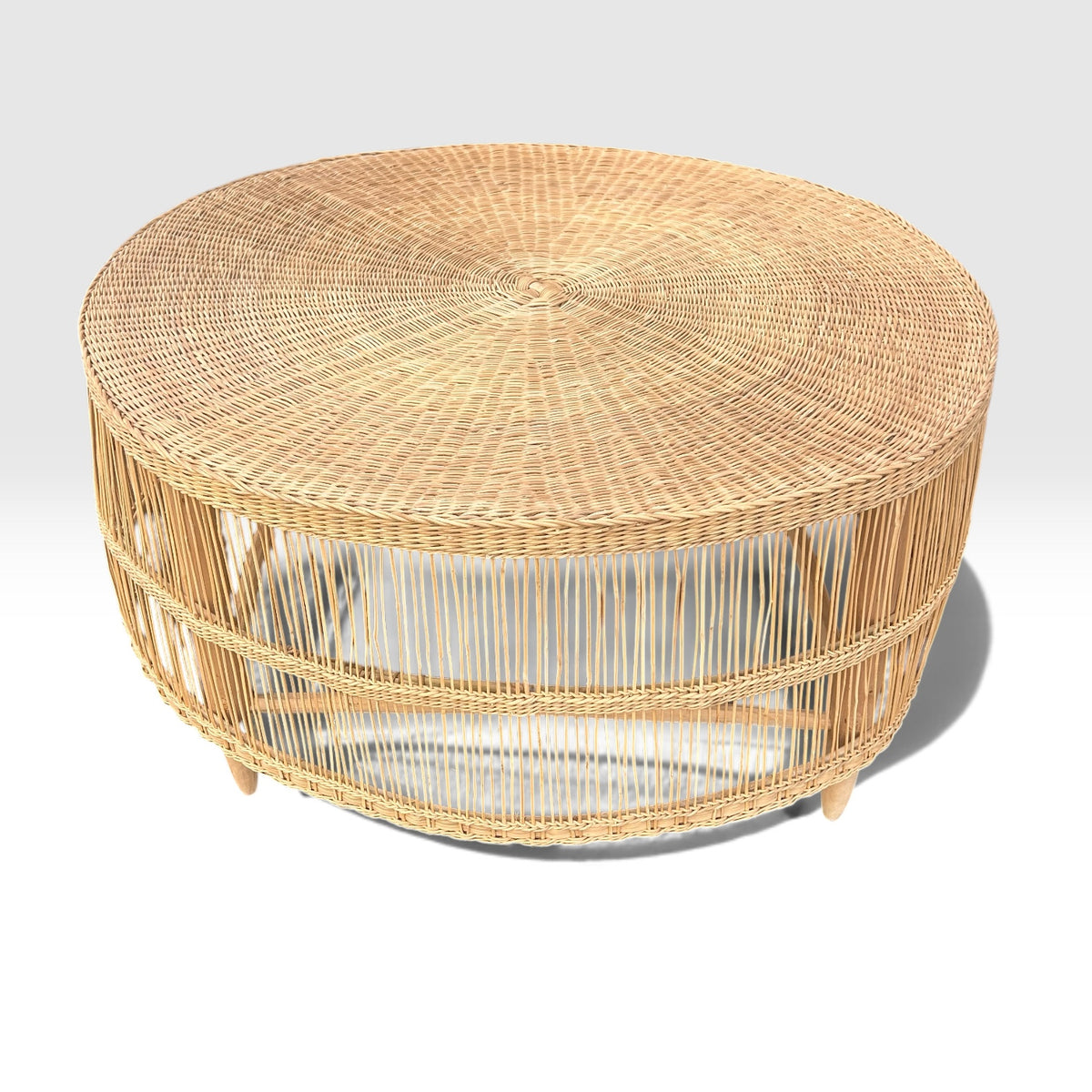 A round woven rattan coffee table with a natural light tan color, featuring a slatted open side design and small legs, shown on a light gray gradient background.