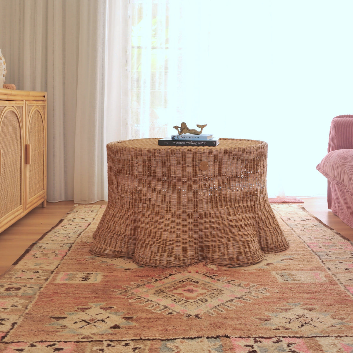 A round woven rattan coffee table draped in a matching natural fiber table skirt, styled in a bright living room on a patterned rug.